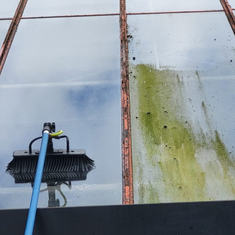 A window cleaner uses a brush and pole to clean a large, dirty window.